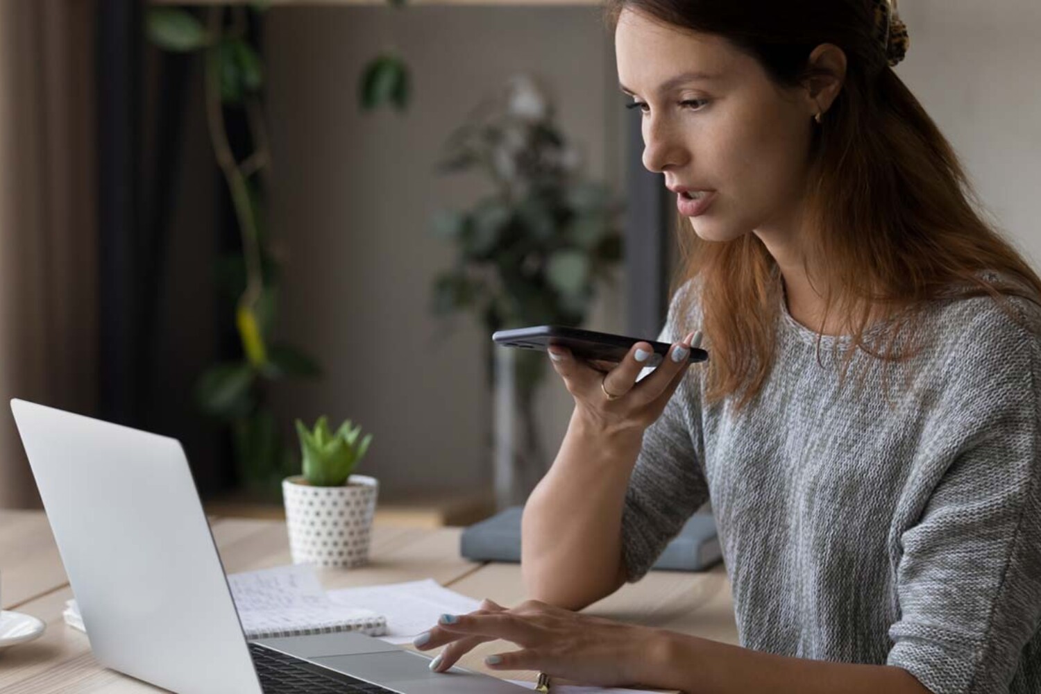 Young woman recording into phone while working on laptop